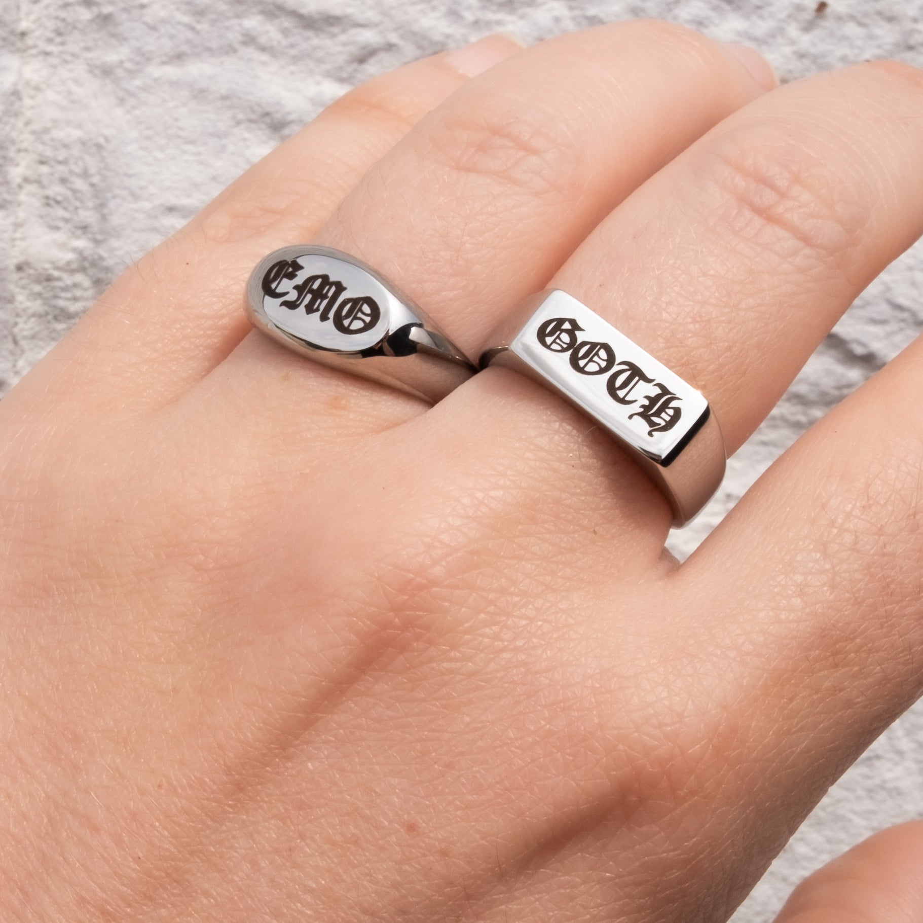 A close-up of a hand wearing two silver rings engraved with religious symbols and text, one ring featuring the word 'GOTH' and the other 'EMO